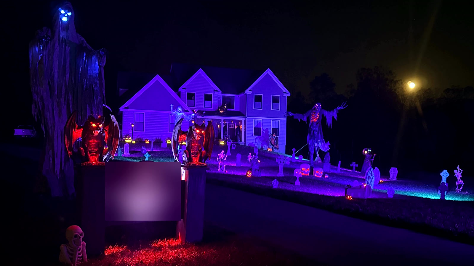 Two-story home washed in purple light with a lawn graveyard, gargoyles and a towering reaper