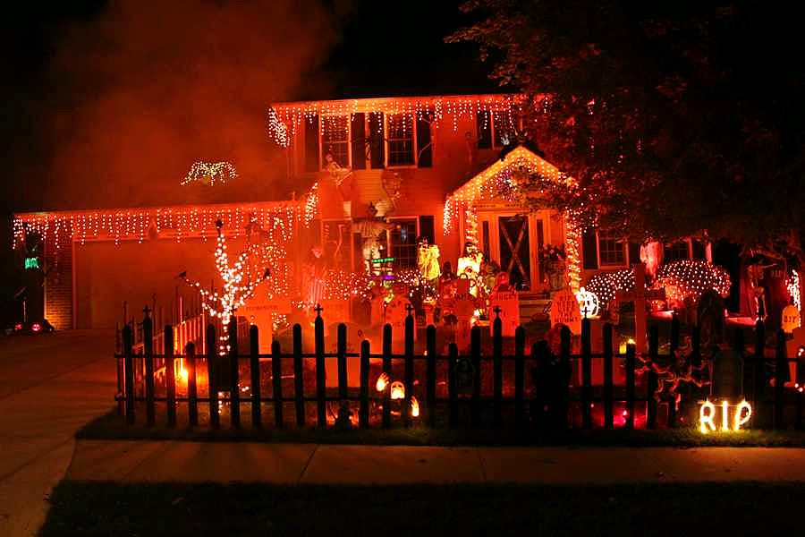 Two-story home glowing orange with roofline lights, a picket fence and tombstones